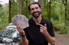 A man holds cash, smiling by a car in a forest.